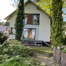 Dom jednorodzinny w Bielsku-Białej z nową, jasną elewacją, widoczny z poziomu ogrodu, z drzewami owiniętymi bluszczem po bokach i metalowym rusztowaniem na pierwszym planie.