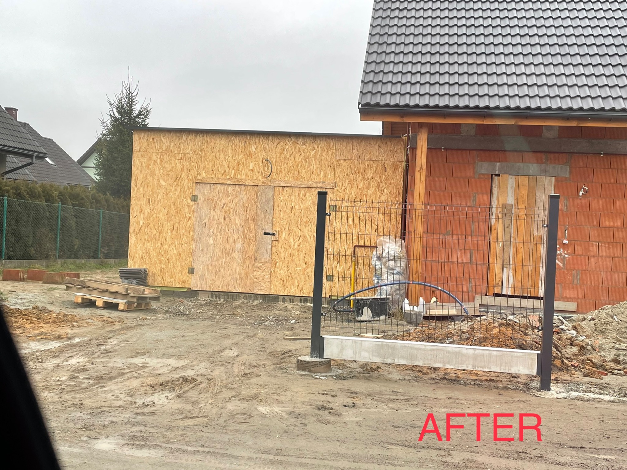 Budowa garażu z płyty OSB obok budynku z czerwonej cegły z zamontowaną bramą wjazdową, widoczne ogrodzenie panelowe i elementy konstrukcyjne, teren wokół w stanie surowym.