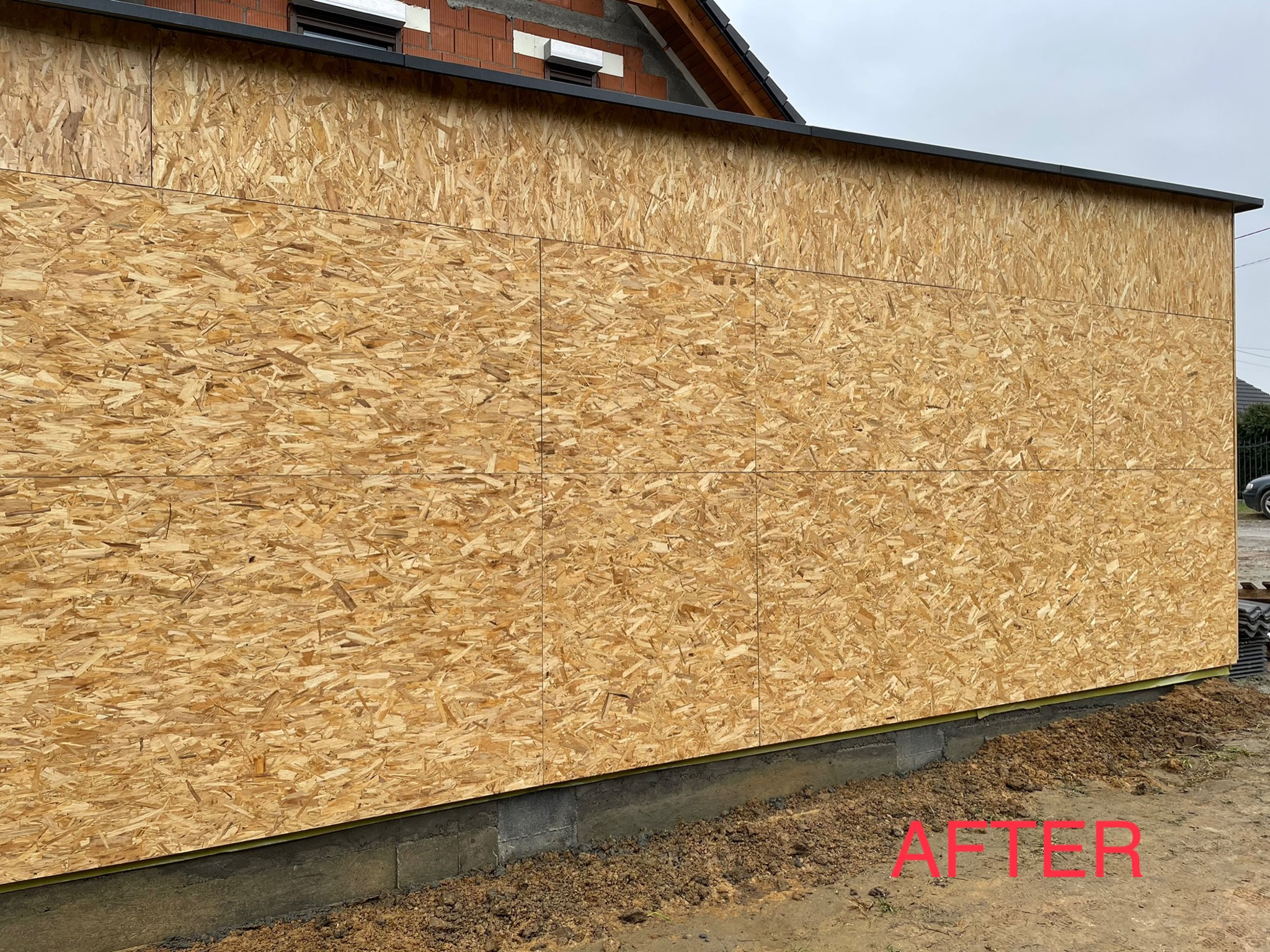 Ściana budynku w trakcie ocieplania, pokryta płytami OSB; widoczny fragment dachu z czarną obróbką blacharską i fragment ściany z cegły.