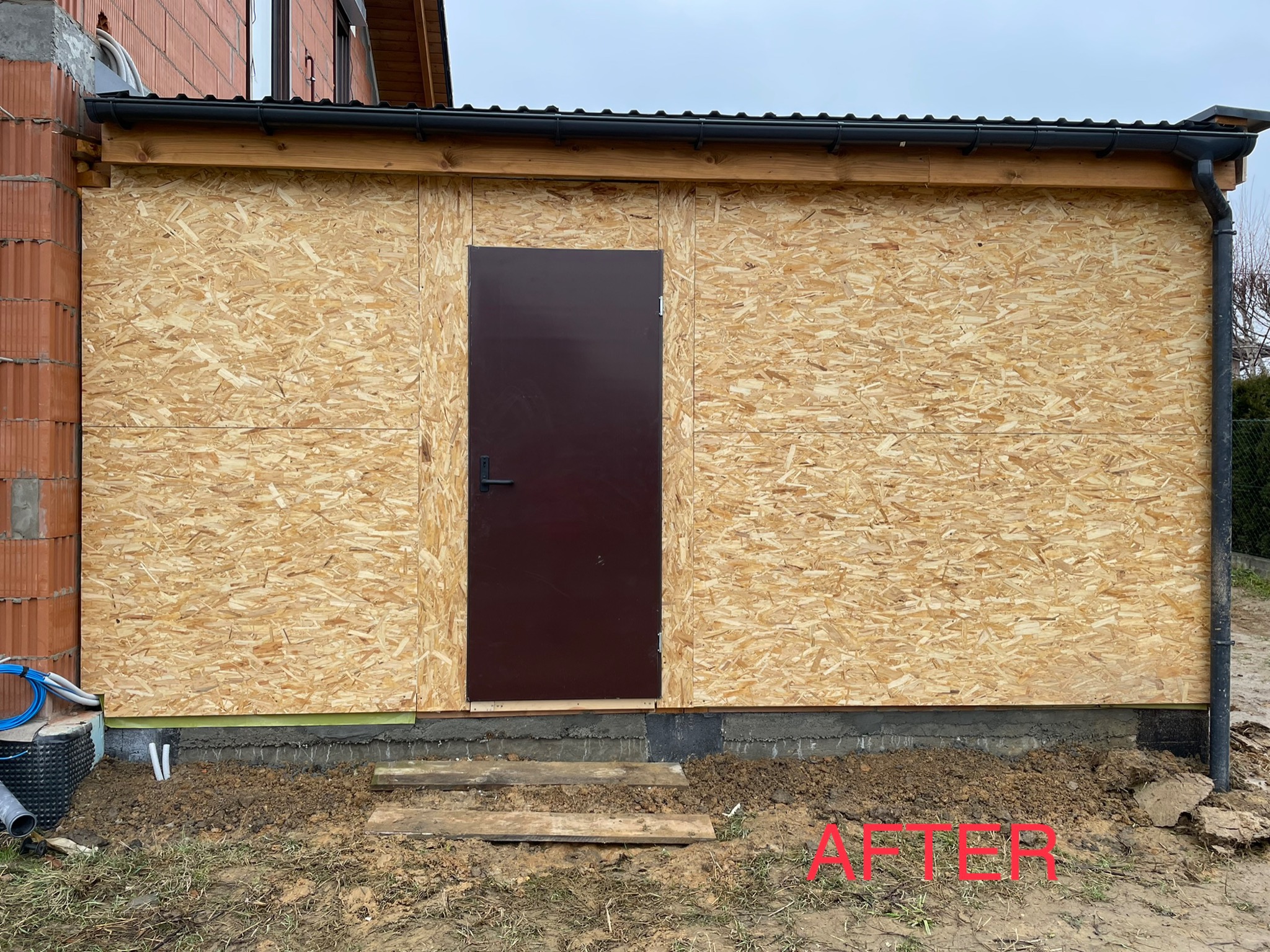 Ściana budynku obłożona płytami OSB z zamontowanymi brązowymi drzwiami wejściowymi, widoczne rynny i fragment dachu, teren wokół budynku częściowo wyłożony drewnianymi deskami.