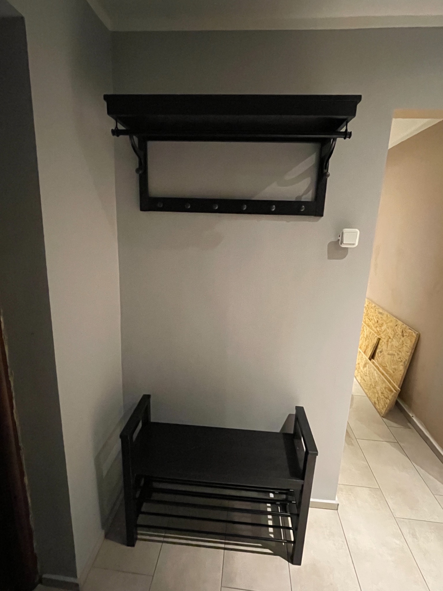 Ciemna szafka na buty z półkami i wieszak z półką zamontowane na szarej ścianie w przedpokoju, widoczna płytka podłogowa i fragment płyty OSB w tle.