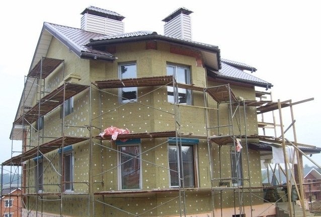 Dwupiętrowy dom w trakcie ocieplania wełną mineralną, zabezpieczoną kołkami, widoczne rusztowania i częściowo odkryta cegła na narożniku budynku.