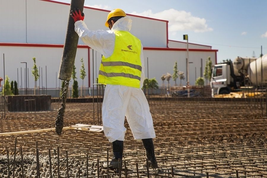 Pracownik w kombinezonie ochronnym i żółtej kamizelce wylewa beton z pompy na zbrojenie fundamentów, w tle budynek przemysłowy i betoniarka.