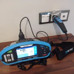 Miernik elektryczny Metrel EurotestXD podłączony do gniazdka elektrycznego z otwartą klapką zabezpieczającą, wyświetlający wyniki pomiarów na ekranie LCD, leżący na drewnianym blacie.