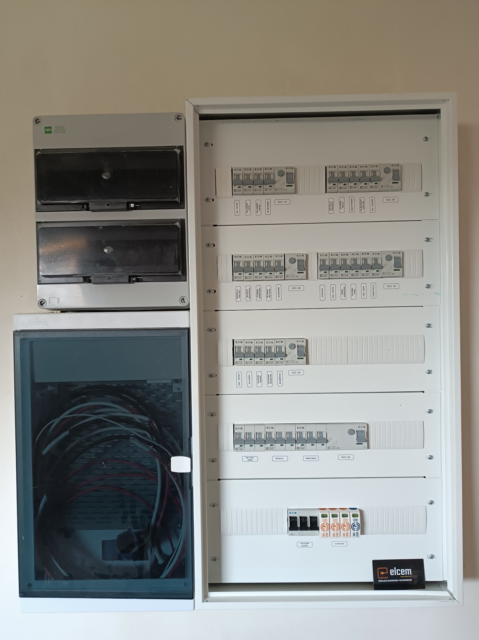 Rozdzielnica elektryczna z zainstalowanymi bezpiecznikami i przewodami w skrzynce zabezpieczającej, widoczne oznaczenia obwodów oraz logo firmy Elcem w prawym dolnym rogu.