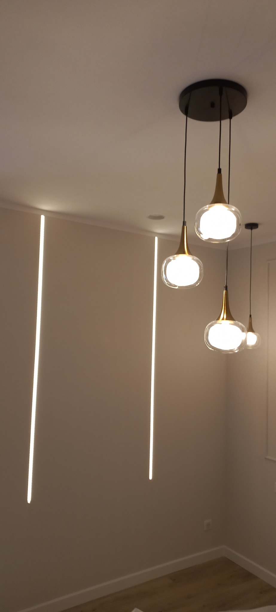 Nowoczesne oświetlenie wnętrza: wiszące lampy z elementami złota i szklanymi kloszami, w tle pionowe pasy świetlne na ścianie.