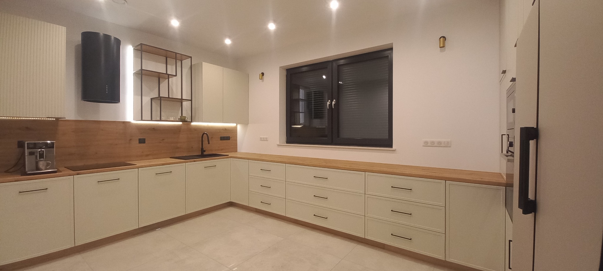 Nowoczesna kuchnia z białymi szafkami, drewnianym blatem i czarnym okapem oraz oświetleniem LED podszafkowym, widoczne gniazdka elektryczne na ścianie.