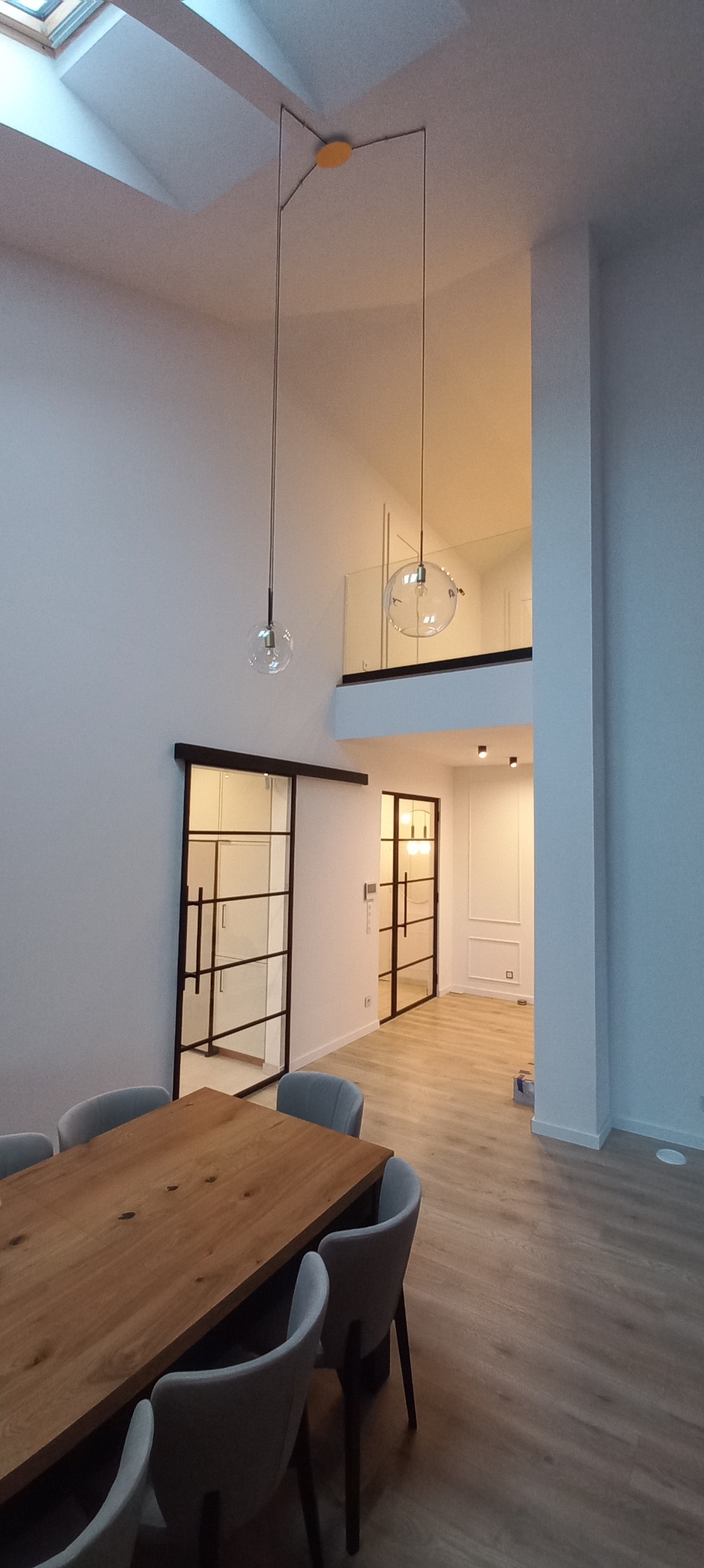Nowoczesne wnętrze z wysokim sufitem, szklanymi kulami jako lampami wiszącymi, drewnianym stołem z szarymi krzesłami, przesuwnymi drzwiami w stylu loft i antresolą z przeszkloną balustradą.