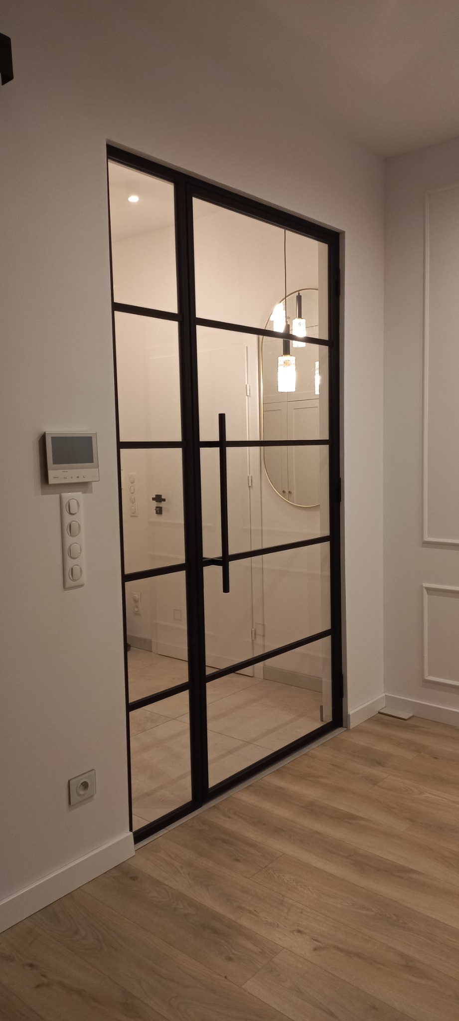 Nowoczesne, przeszklone drzwi loftowe z czarnymi szprosami, zamontowane w białej ścianie, widoczny panel sterowania inteligentnego domu i wykończenie podłogi panelami.