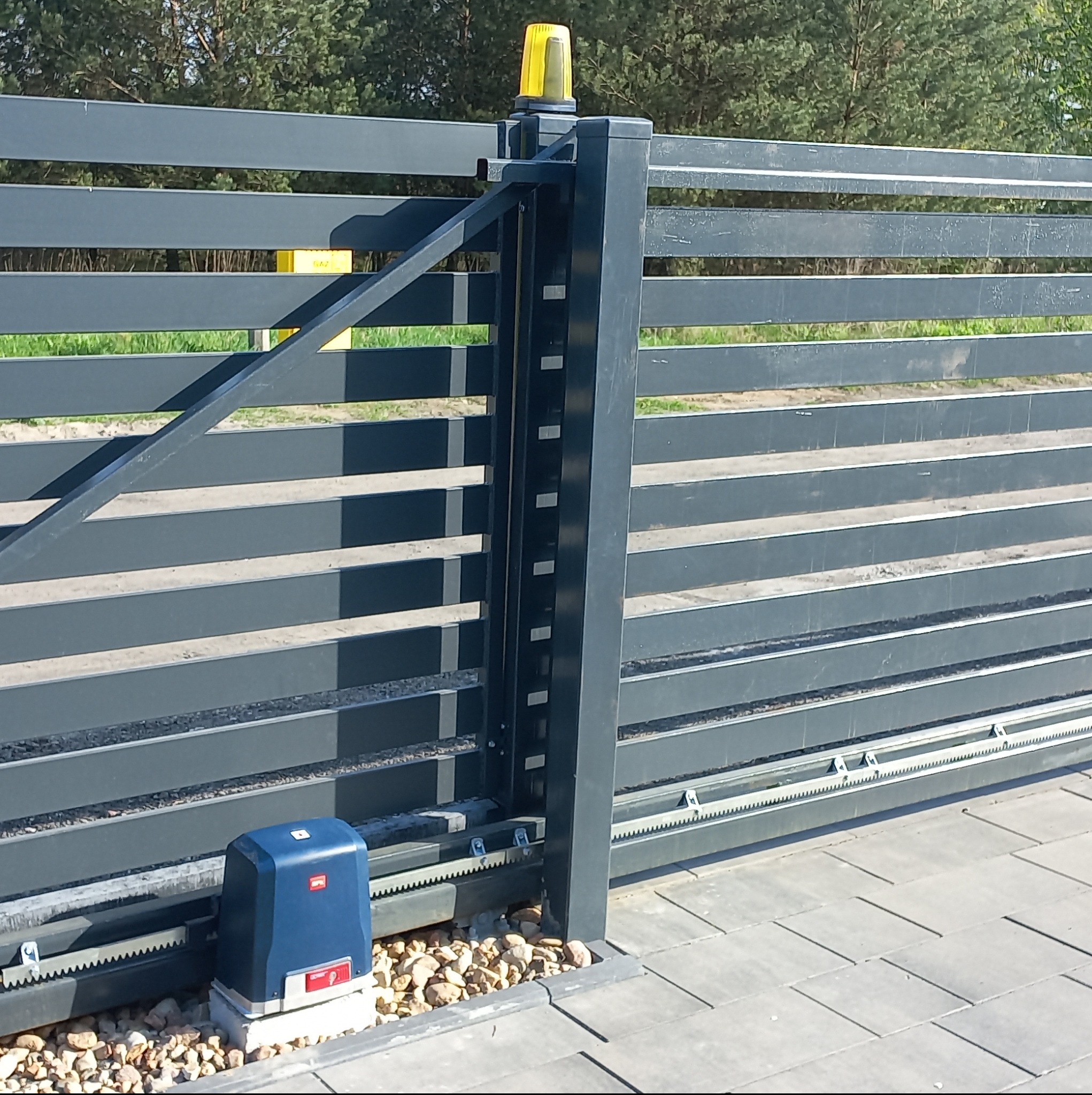 Automatyczna brama przesuwna z napędem i lampą ostrzegawczą, widoczna szyna z zębatką i kostka brukowa u podstawy.