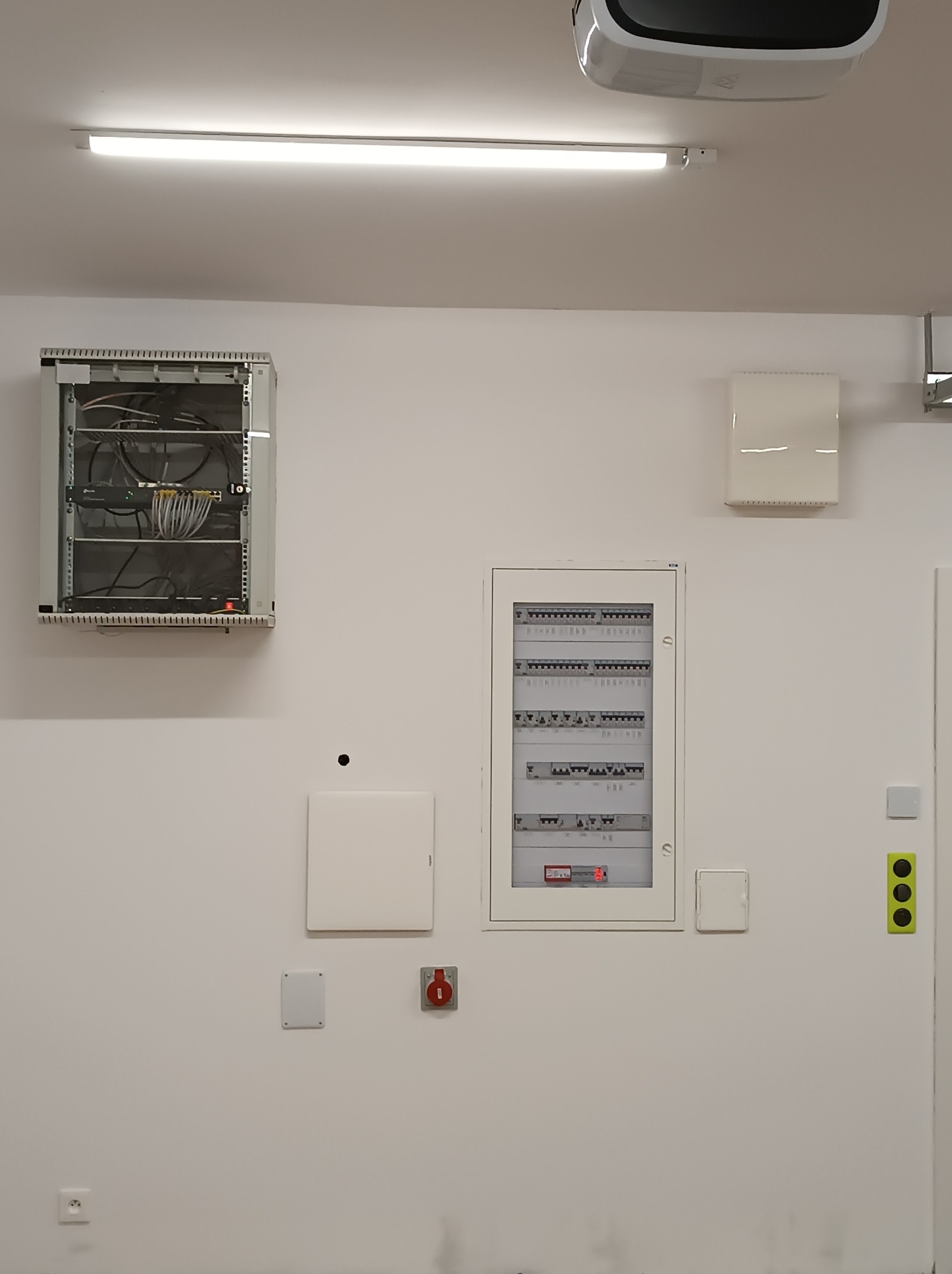 Rozdzielnica elektryczna z zabezpieczeniami, szafa teleinformatyczna z patch panelem, przyciski sterujące oświetleniem i gniazdko elektryczne na białej ścianie w pomieszczeniu z oświetleniem...