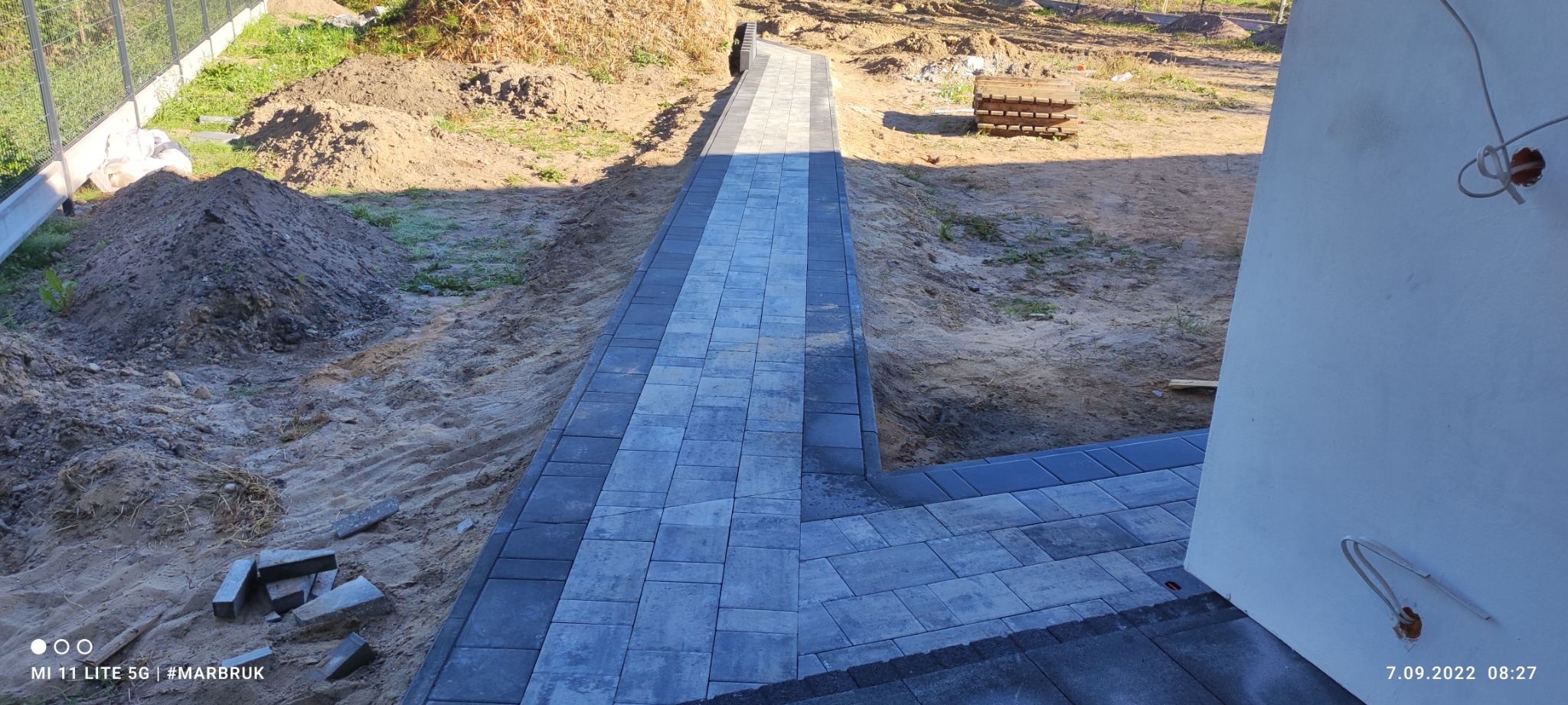 Ścieżka z szarej kostki brukowej prowadząca wzdłuż budynku w trakcie budowy, widoczne przygotowane podłoże i niezakończone prace.