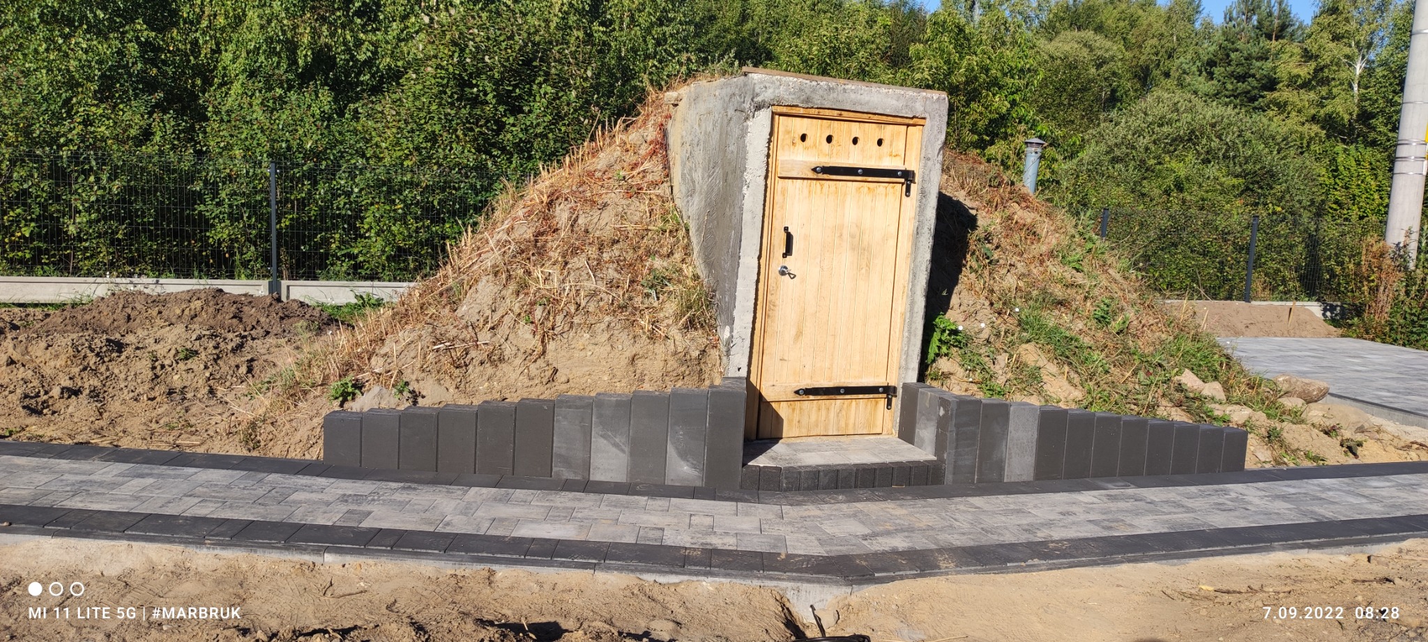 Zakończona budowa ziemianki z drewnianymi drzwiami, otoczonej nową ścieżką z szarej kostki brukowej i obrzeżami z ciemnych bloczków betonowych na tle zieleni.