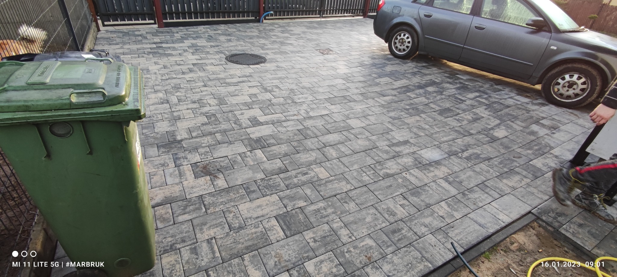Ułożona szara kostka brukowa na podjeździe, widoczny fragment samochodu i kosz na śmieci, zdjęcie zrobione z niskiej perspektywy.