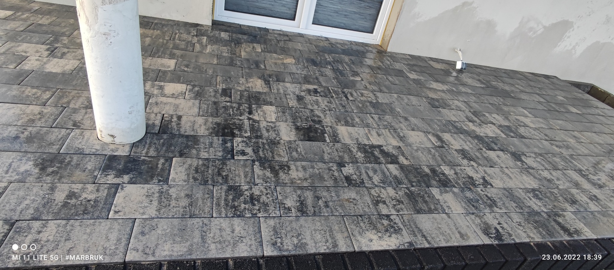 Ułożona kostka brukowa w odcieniach szarości i beżu, z widocznym słupem i fragmentem okna w tle, wykonana z podłużnych elementów o nieregularnej fakturze.