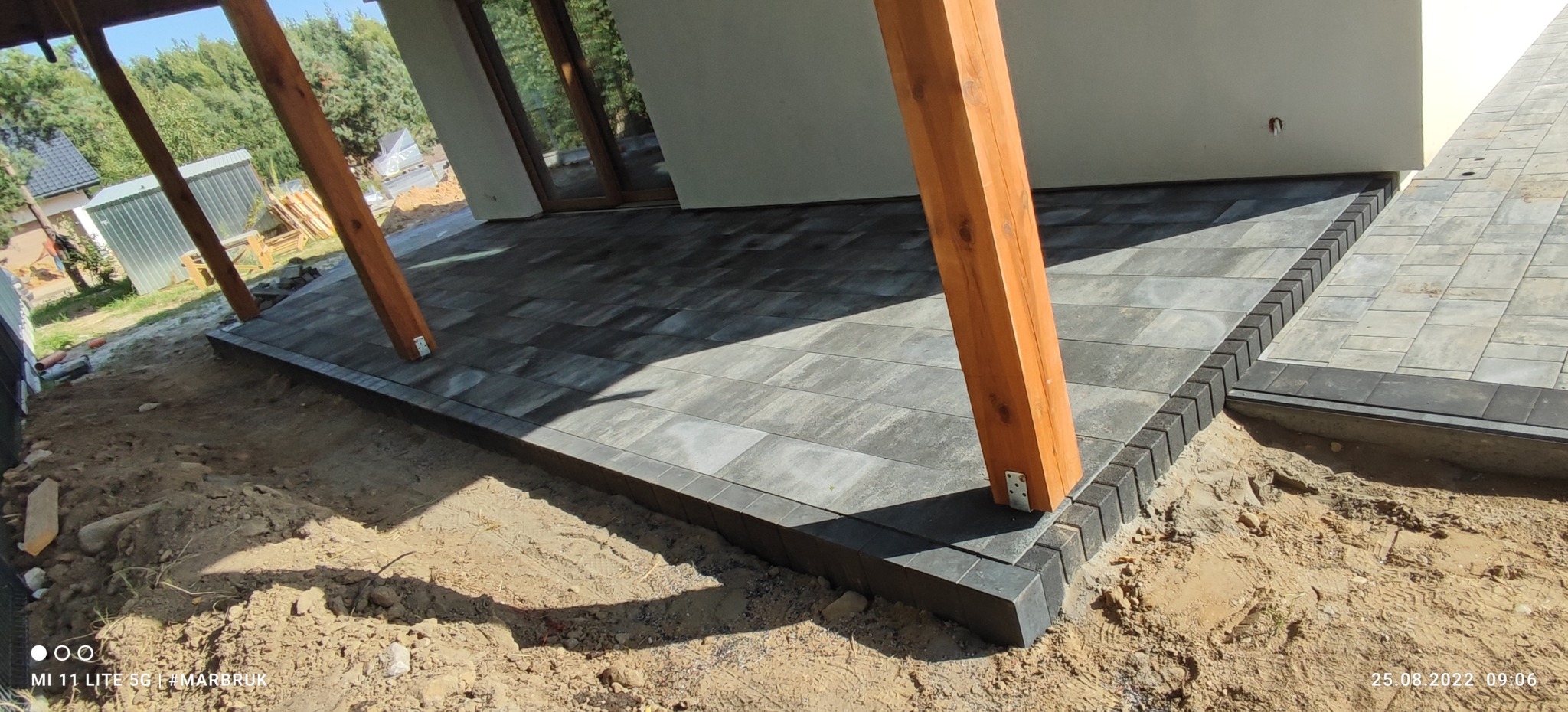 Nowoczesny taras z szarej kostki brukowej ułożonej przy jasnym budynku z drewnianymi słupami podtrzymującymi zadaszenie, widoczne obrzeża i podsypka piaskowa.