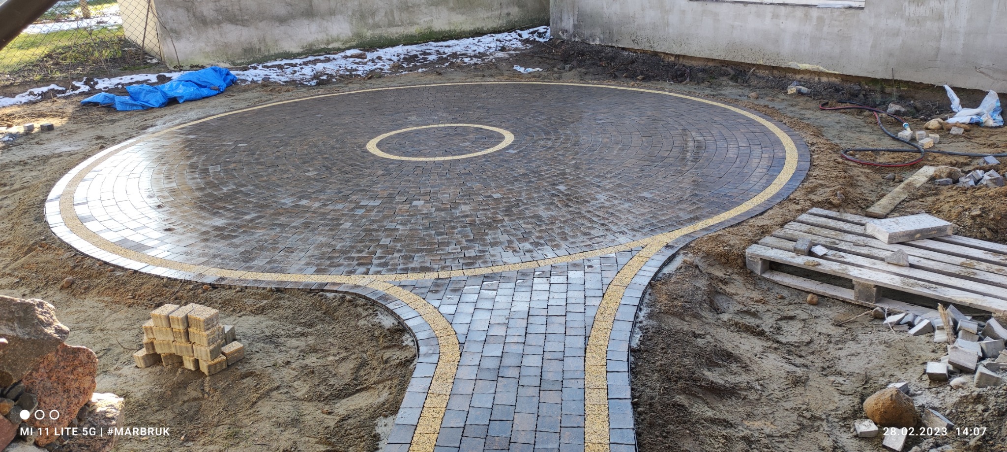 Okrągły plac z ciemnej kostki brukowej z żółtą obwódką i ścieżką, w tle budynek i resztki śniegu, widoczne narzędzia i materiały budowlane.