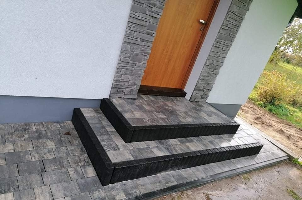 Schody wejściowe do domu wyłożone kostką brukową w odcieniach szarości, z ciemnym obramowaniem, pasujące do elewacji z elementami kamiennymi.
