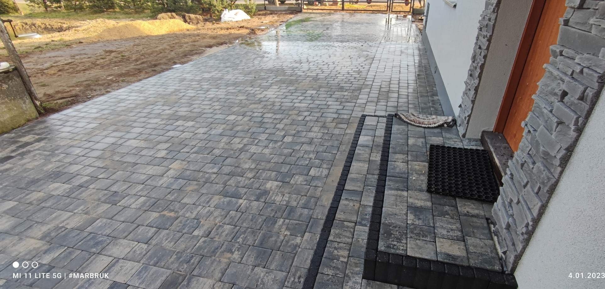 Ułożona szara kostka brukowa prowadząca do wejścia domu z kamienną elewacją, widoczne wycieraczki przy drzwiach wejściowych.