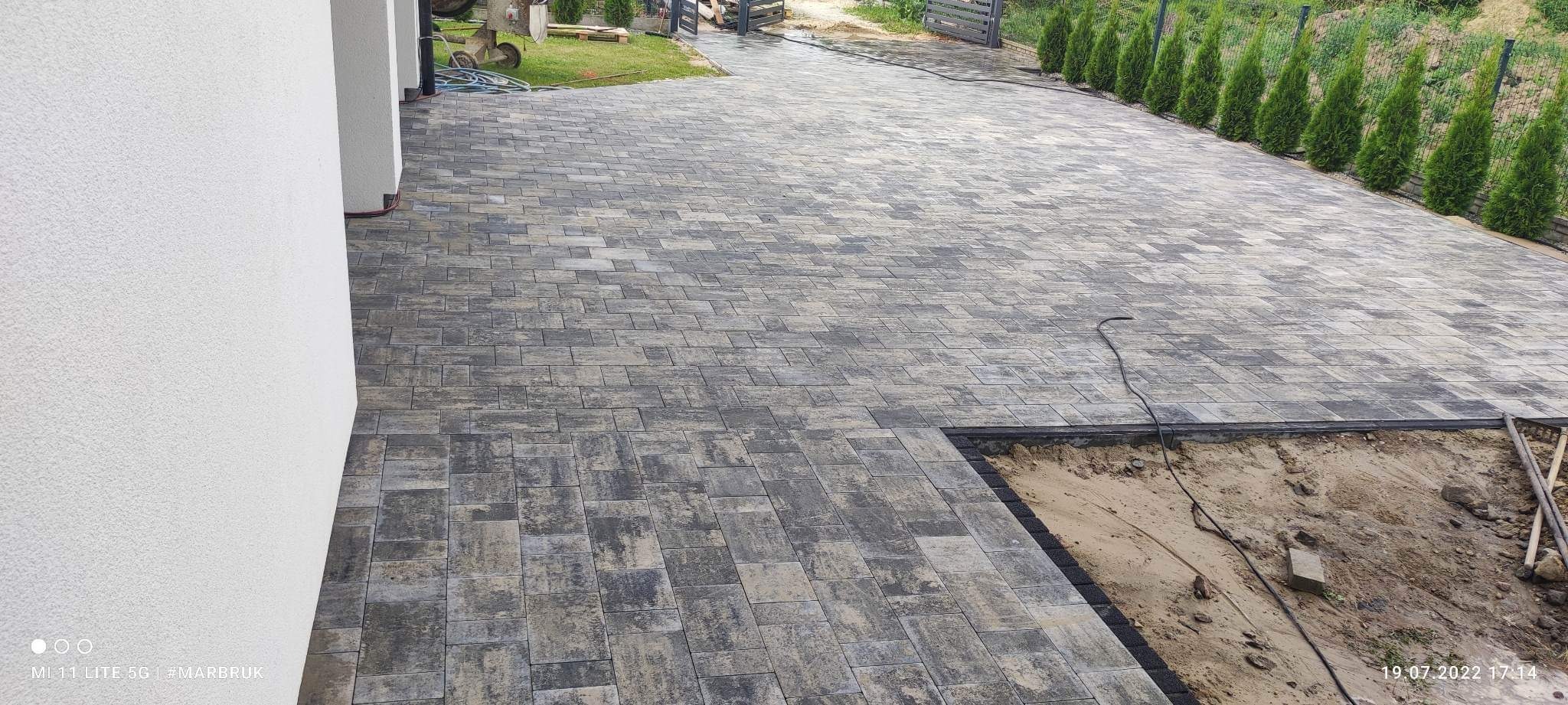 Nowo ułożona, wielobarwna kostka brukowa na podjeździe, z widocznym fragmentem przygotowywanego podłoża i zielonymi tujami w tle.