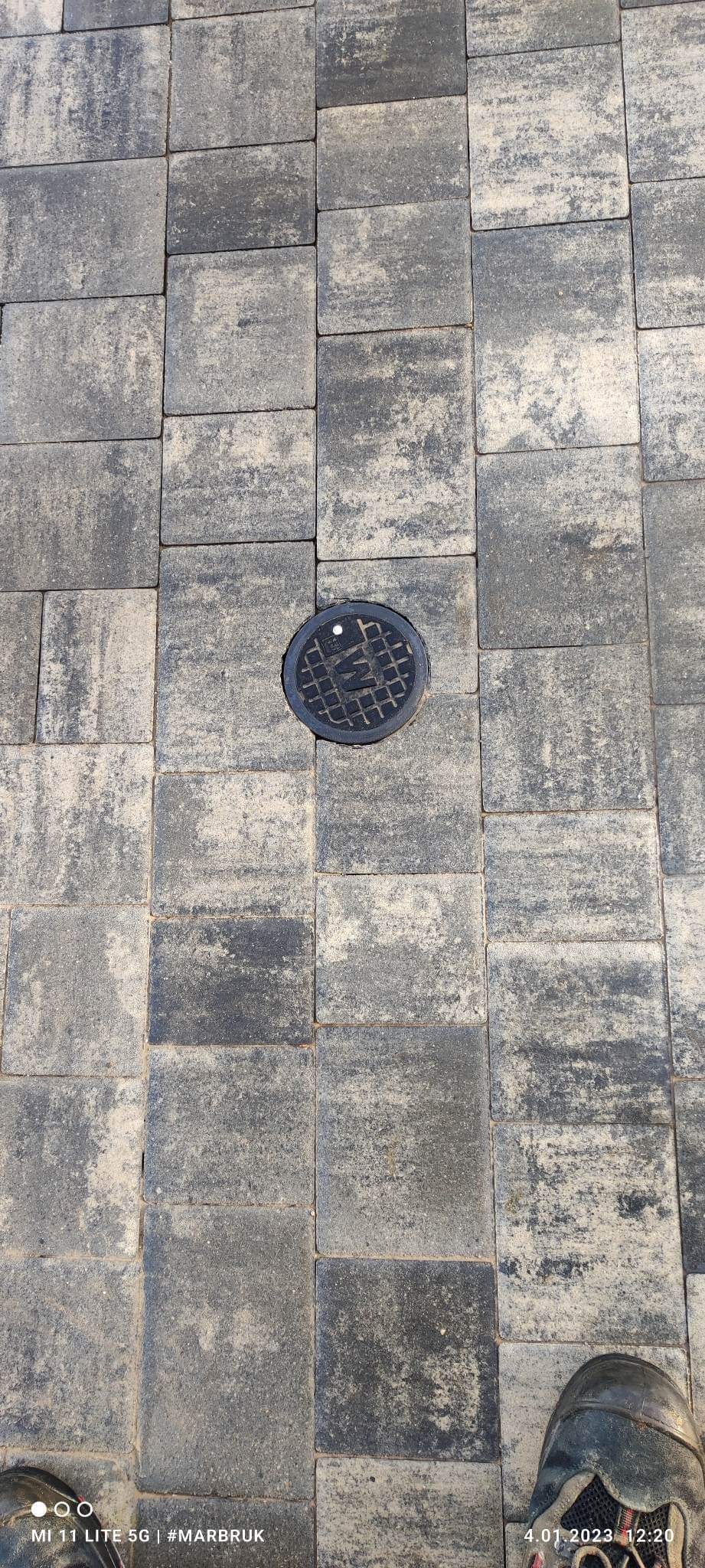 Szare, kwadratowe kostki brukowe ułożone wokół okrągłej studzienki kanalizacyjnej, widok z góry, fragment buta w prawym dolnym rogu.