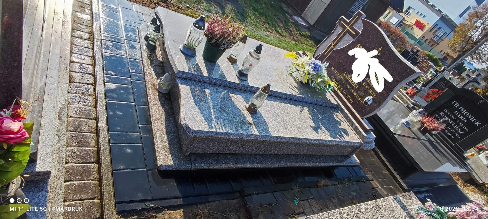 Ciemna kostka brukowa ułożona wokół granitowego nagrobka z zapalonymi zniczami i kwiatami, widok z góry.