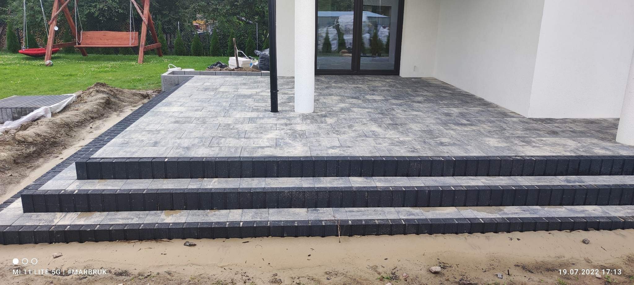 Schody wejściowe do domu wyłożone szarą kostką brukową z ciemnoszarą obwódką, widok z dołu na trzystopniowe schody i fragment tarasu.