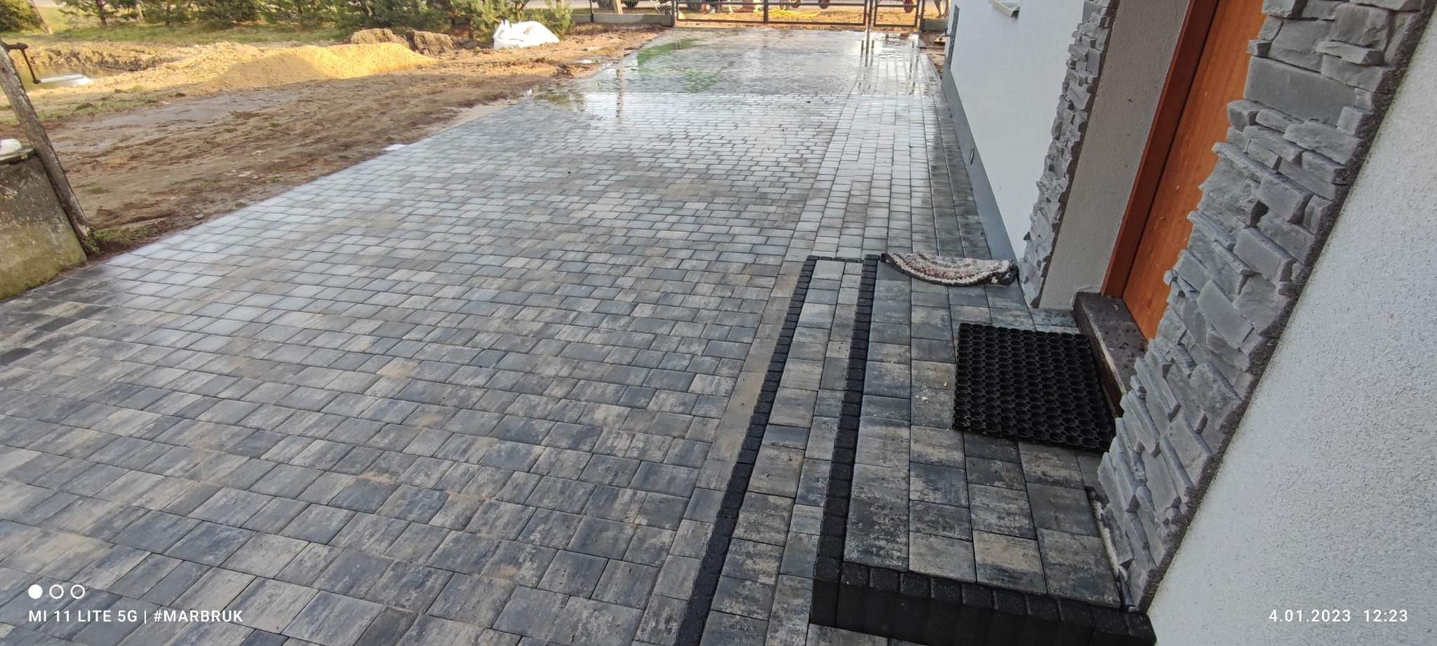 Nowo ułożona, szara kostka brukowa prowadząca do wejścia domu, z widocznym dywanikiem i wykończeniem elewacji imitującym kamień.