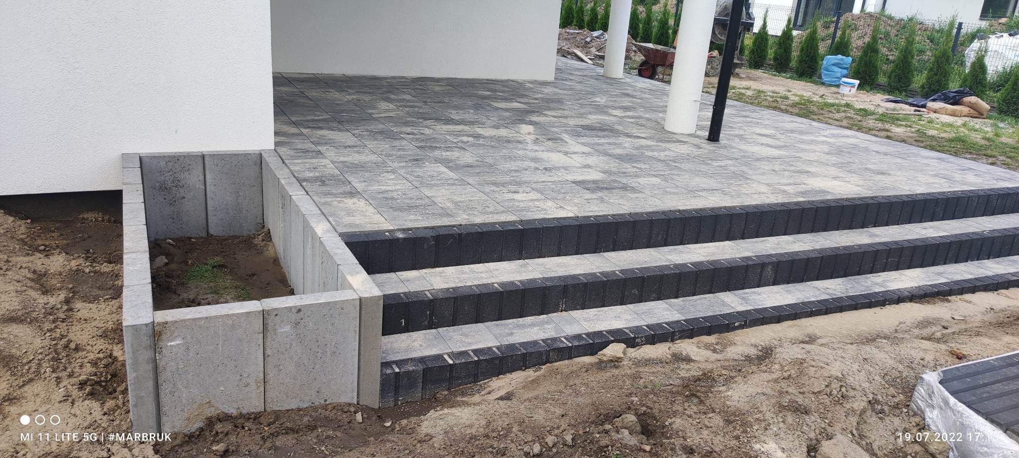 Wykończony taras z szarej kostki brukowej, z trzema stopniami obłożonymi ciemną kostką, widoczna ziemia i pozostałości po budowie.