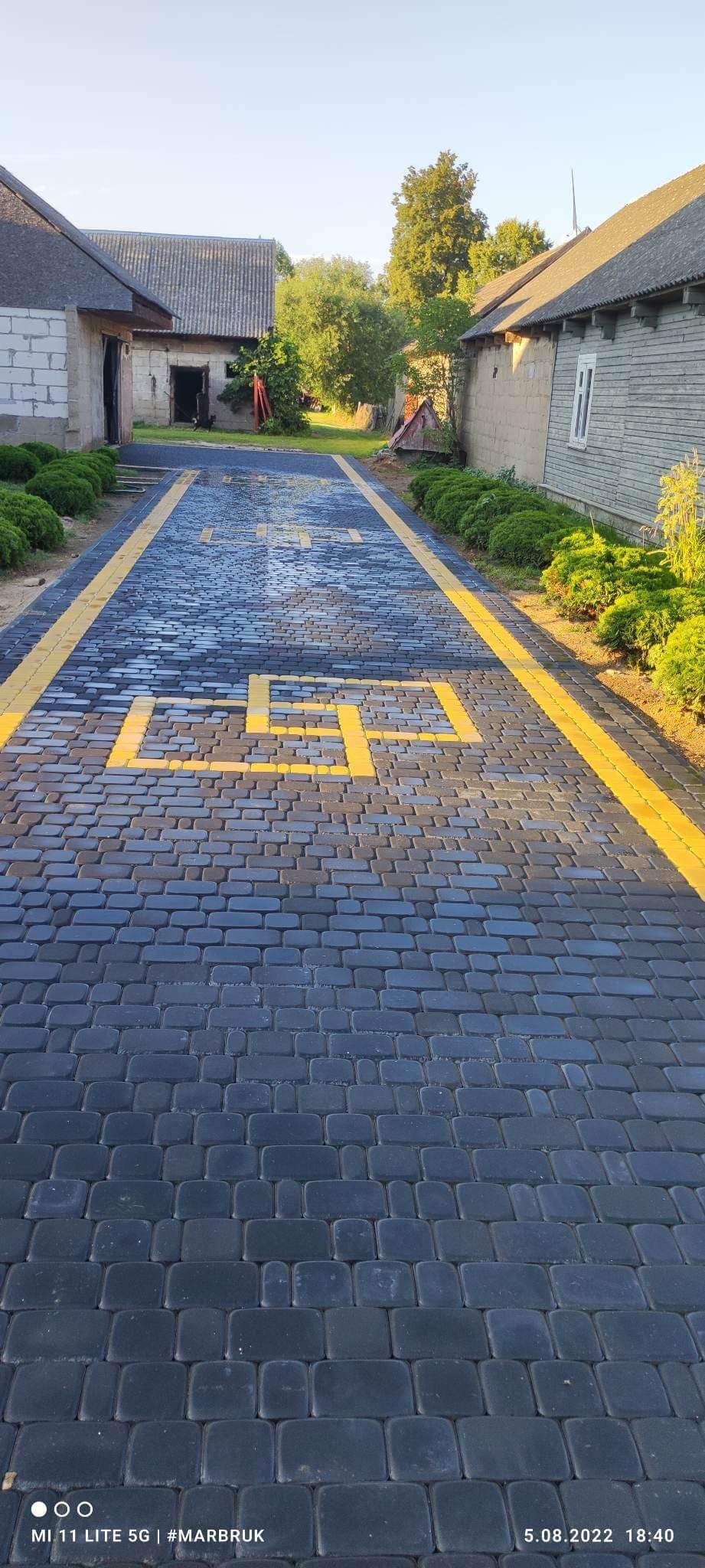 Ułożona nawierzchnia z ciemnej kostki brukowej z żółtymi liniami i znakami poziomymi, prowadząca do budynków gospodarczych w wiejskim otoczeniu.