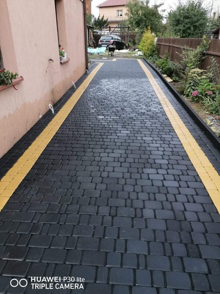 Ułożona, ciemnoszara kostka brukowa z żółtymi obrzeżami, prowadząca wzdłuż budynku w kierunku zaparkowanego samochodu i grilla.
