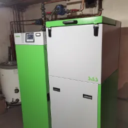 Nowoczesny piec na paliwo stałe SAS Bio Compact z zielonym panelem bocznym i białym korpusem, widoczne rury instalacji grzewczej na ścianie w tle.