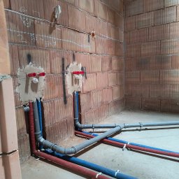 PW Dobry Klimat - Wewnętrzne instalacje wodne i kanalizacyjne w trakcie montażu w surowym pomieszczeniu z ceglanymi ścianami, widoczne rury w kolorach czerwonym, niebieskim i szarym, mocowane do ściany.