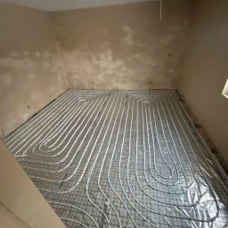 PW Dobry Klimat - Białe rurki ogrzewania podłogowego ułożone na metalowej siatce na folii izolacyjnej w surowym pomieszczeniu przed wylewką.