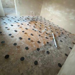 PW Dobry Klimat - Przygotowanie podłoża pod ogrzewanie podłogowe z widocznymi czarnymi podkładkami poziomującymi rozmieszczonymi na surowej wylewce, poziomica leżąca obok, surowe ściany w tle.