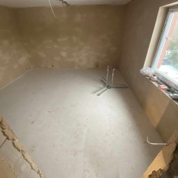 PW Dobry Klimat - Surowe wnętrze w trakcie remontu: świeżo wylana wylewka podłogowa, tynkowane ściany z widocznymi niedoskonałościami, metalowy stojak na środku pomieszczenia, okno z widocznymi elementami...