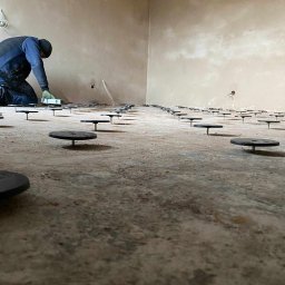 PW Dobry Klimat - Instalacja ogrzewania podłogowego: widok z niskiego kąta na przygotowany beton z rozmieszczonymi wspornikami poziomującymi, instalator z poziomicą w tle.