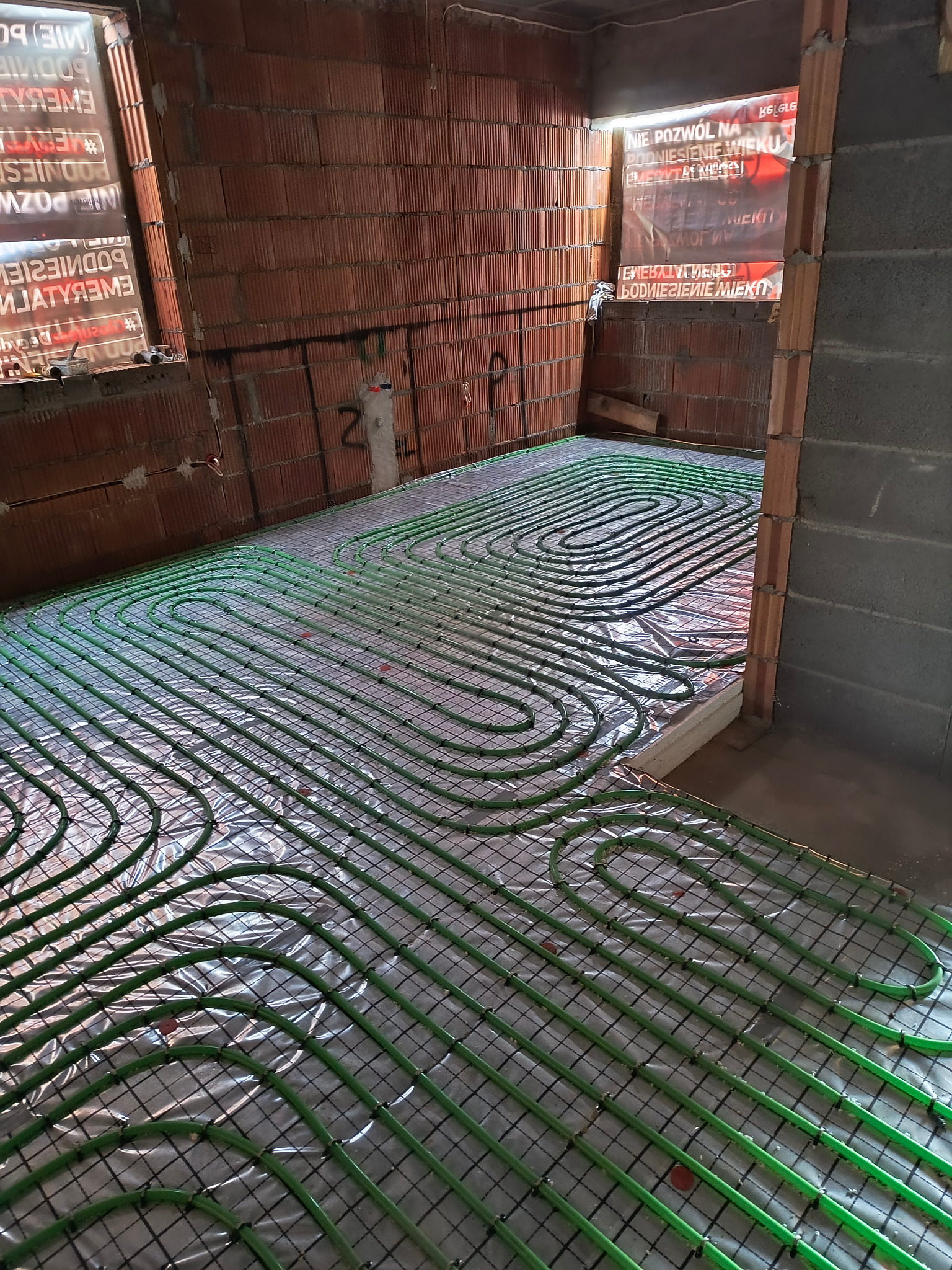 Wnętrze w trakcie instalacji ogrzewania podłogowego z widocznymi zielonymi rurami ułożonymi na folii i siatce, surowe ściany z cegły i betonu w tle.