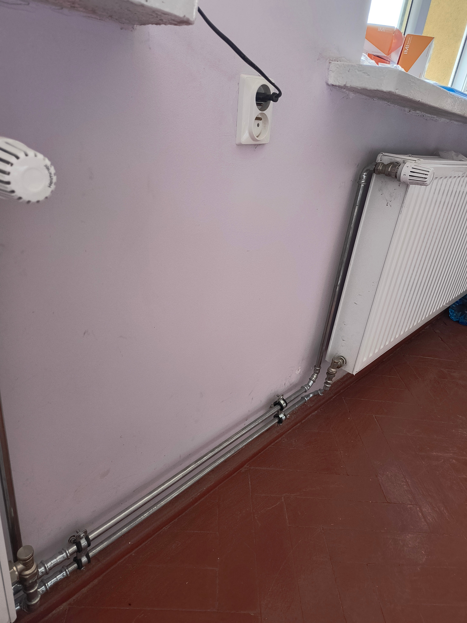 Biały grzejnik żeberkowy z termostatem, podłączony rurami do instalacji grzewczej w pomieszczeniu w Gliwicach, widoczne mocowania rur do ściany i gniazdko elektryczne powyżej.