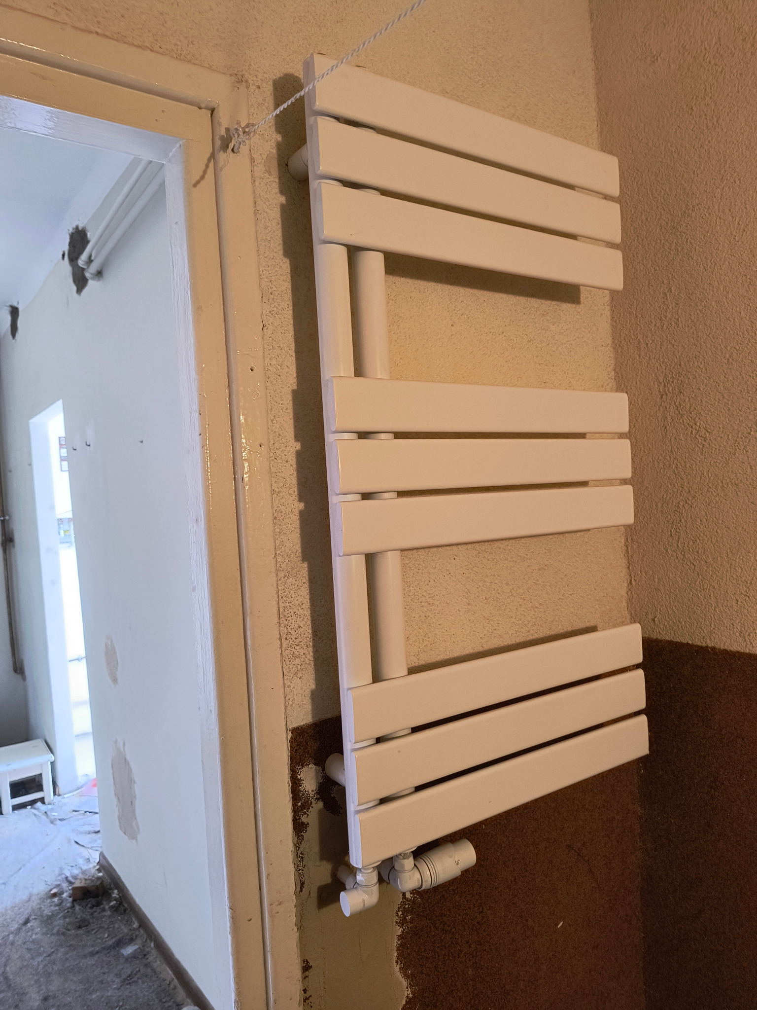 Biały, drabinkowy grzejnik łazienkowy zamontowany na ścianie w trakcie remontu, widoczne niedoskonałości ścian i podłogi.