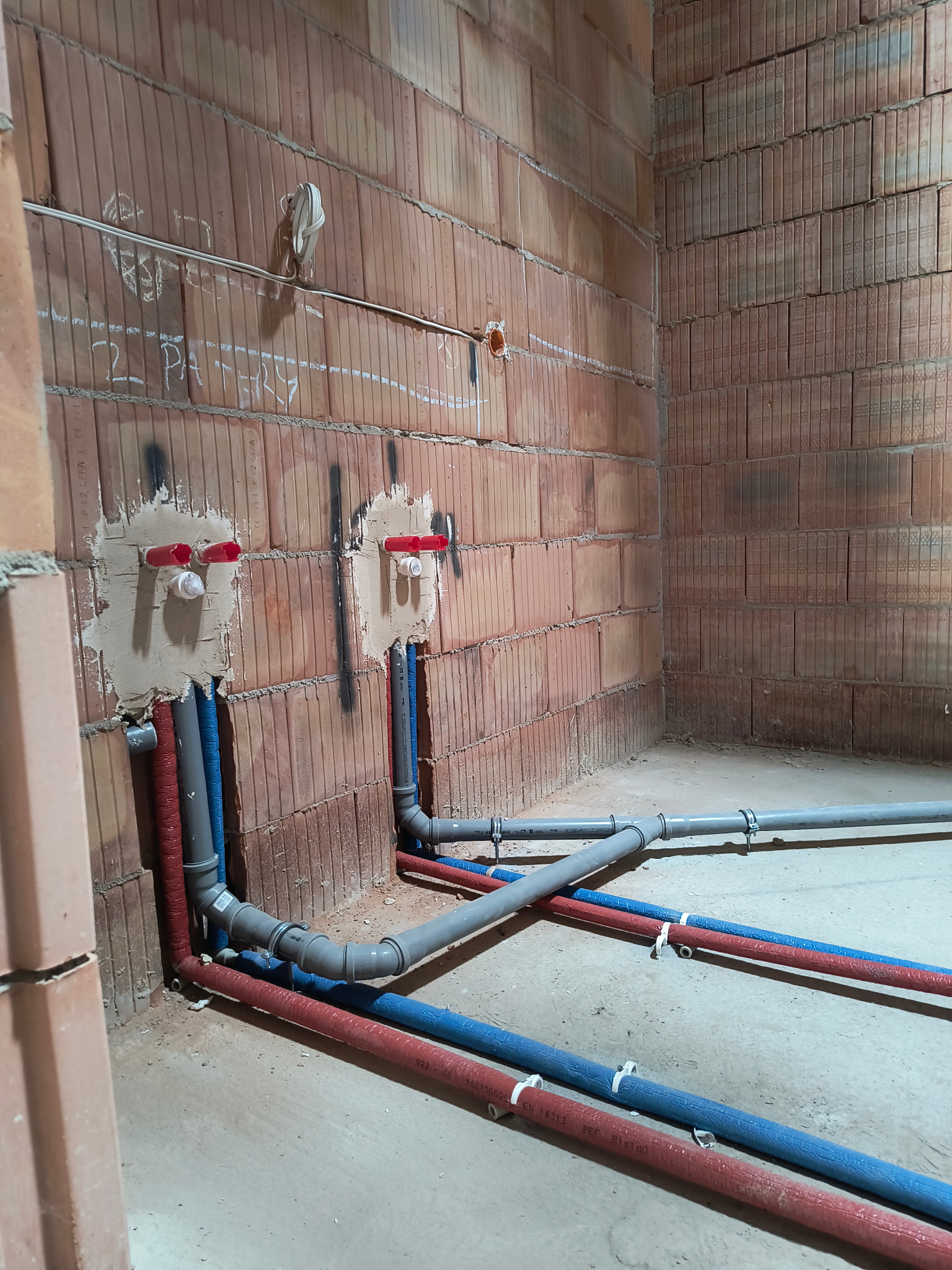 Wewnętrzne instalacje wodne i kanalizacyjne w trakcie montażu w surowym pomieszczeniu z ceglanymi ścianami, widoczne rury w kolorach czerwonym, niebieskim i szarym, mocowane do ściany.
