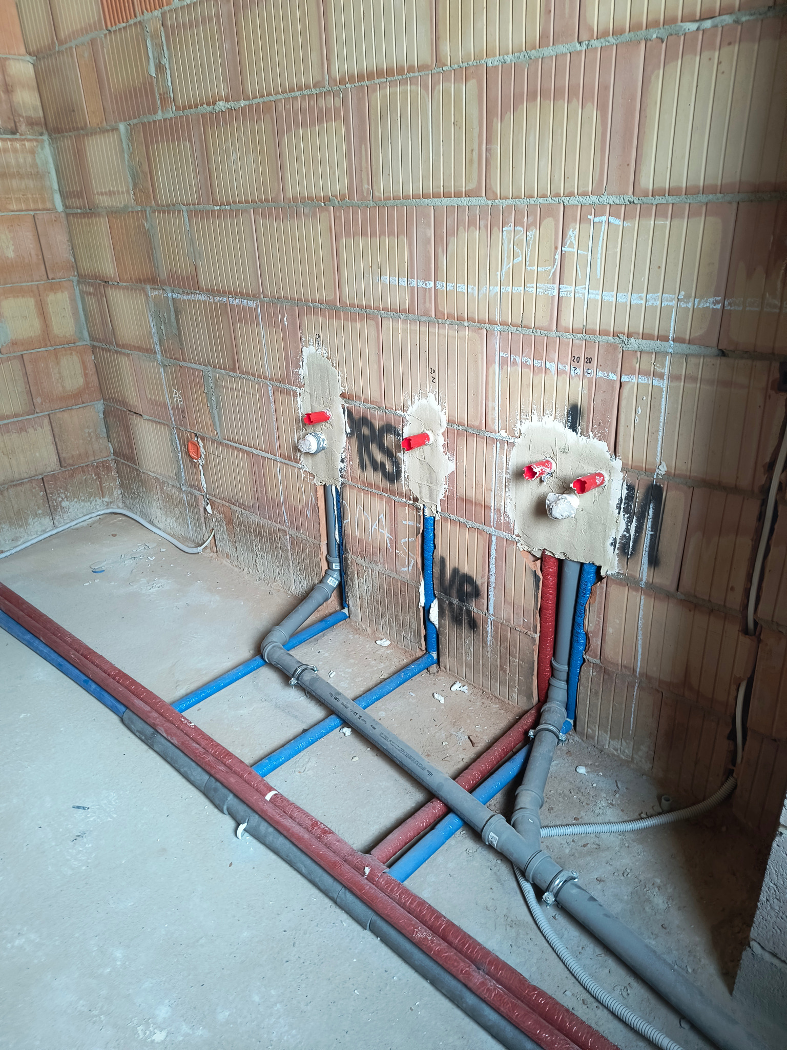 Instalacja wodna w surowym stanie: rury niebieskie i czerwone prowadzone po podłodze i w ścianie z cegły, z zamontowanymi zaworami, częściowo zakryte tynkiem.