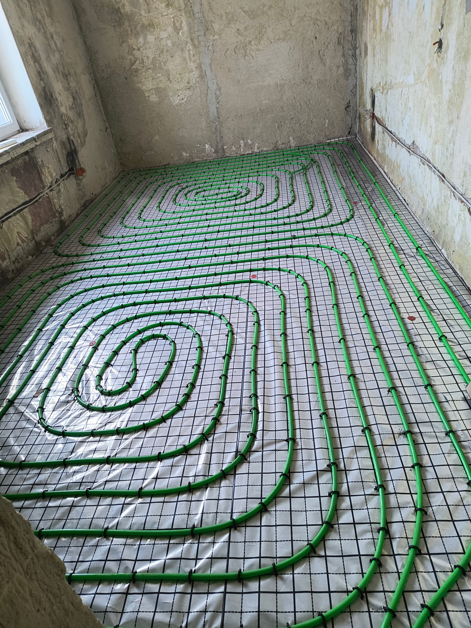 Zielone rury ogrzewania podłogowego ułożone spiralnie na czarnej siatce z białym podkładem, surowy stan wykończenia wnętrza.