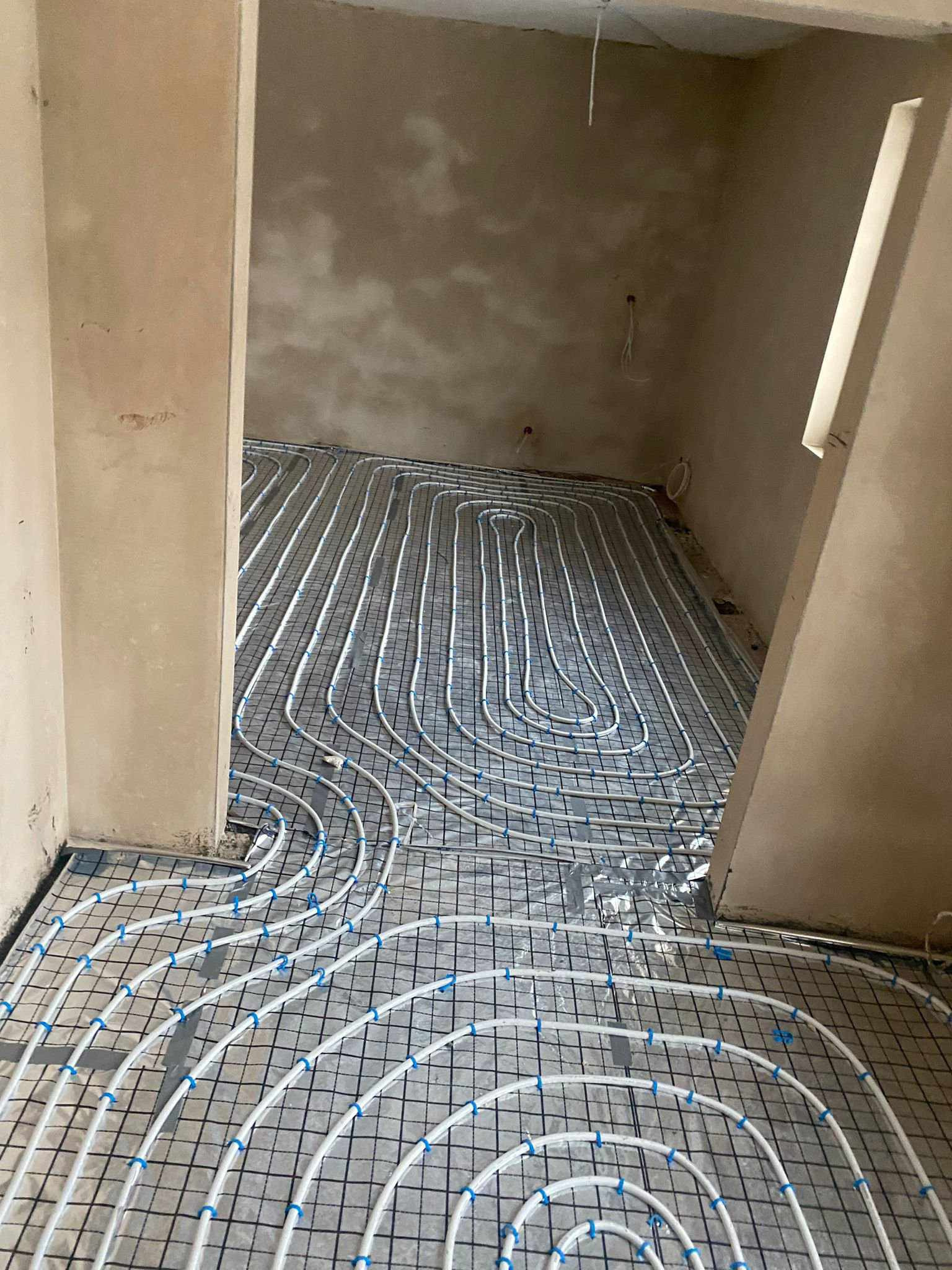 Białe rury ogrzewania podłogowego ułożone na czarnej siatce w surowym pomieszczeniu, widok z perspektywy wejścia, jasne światło.