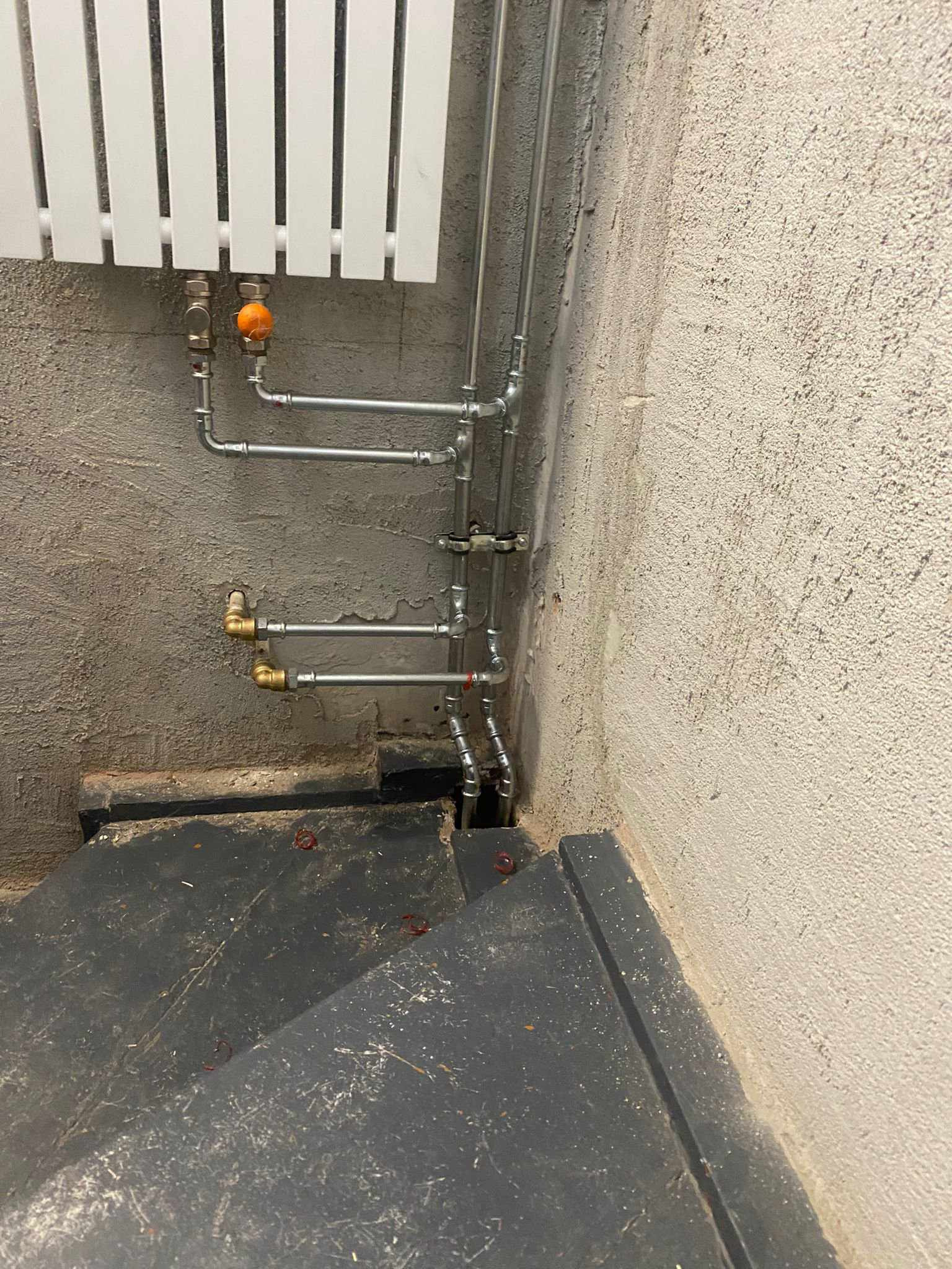 Montaż grzejnika panelowego z instalacją hydrauliczną na ścianie z surowym tynkiem, widoczne rury miedziane i zawory, instalacja częściowo ukryta za schodami.