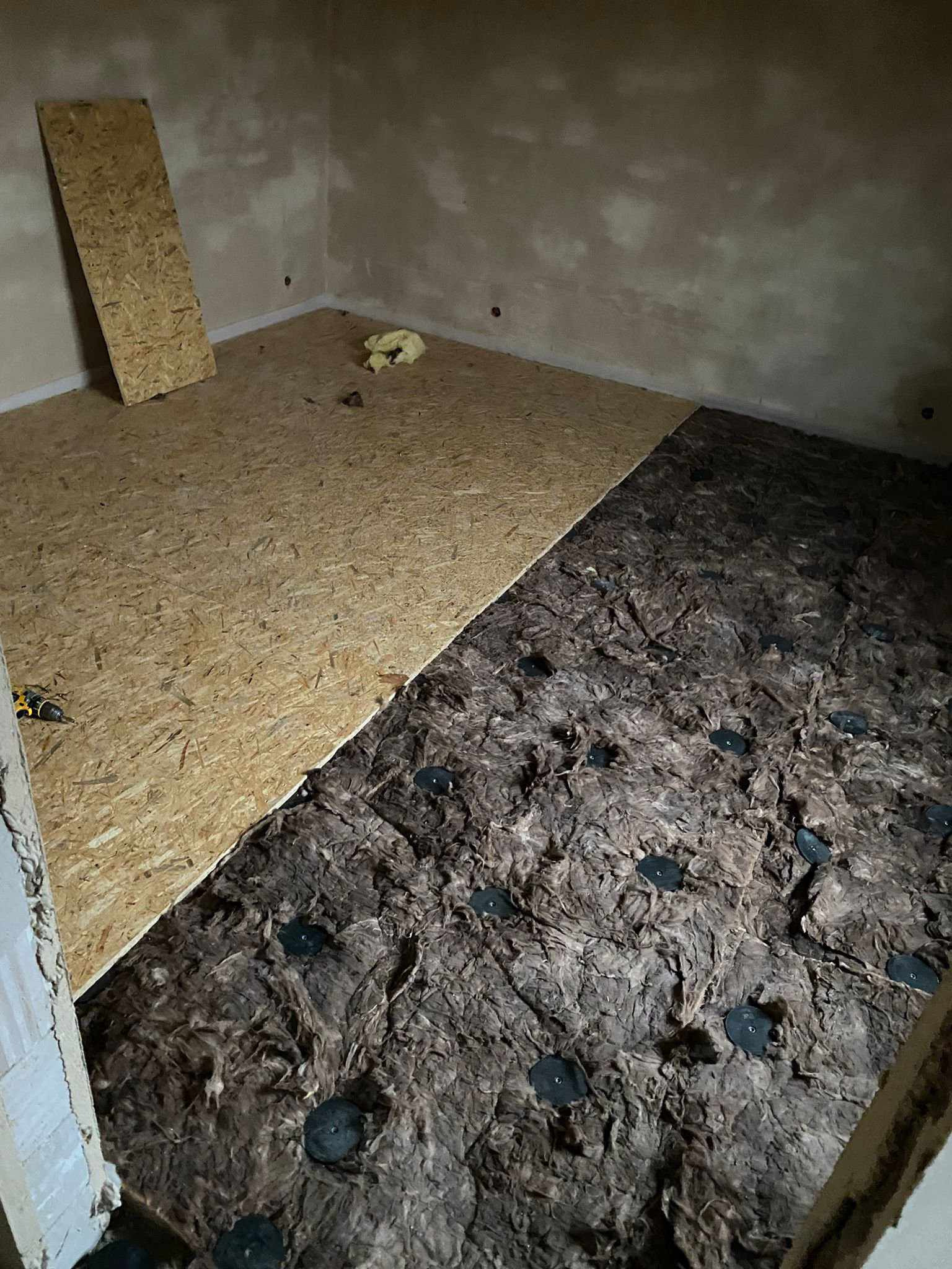 Fragment pokoju w trakcie prac wykończeniowych: część podłogi pokryta izolacją z wełny mineralnej z czarnymi mocowaniami, druga część wyłożona płytą OSB, w rogu pokoju oparta płyta OSB, widoczny...