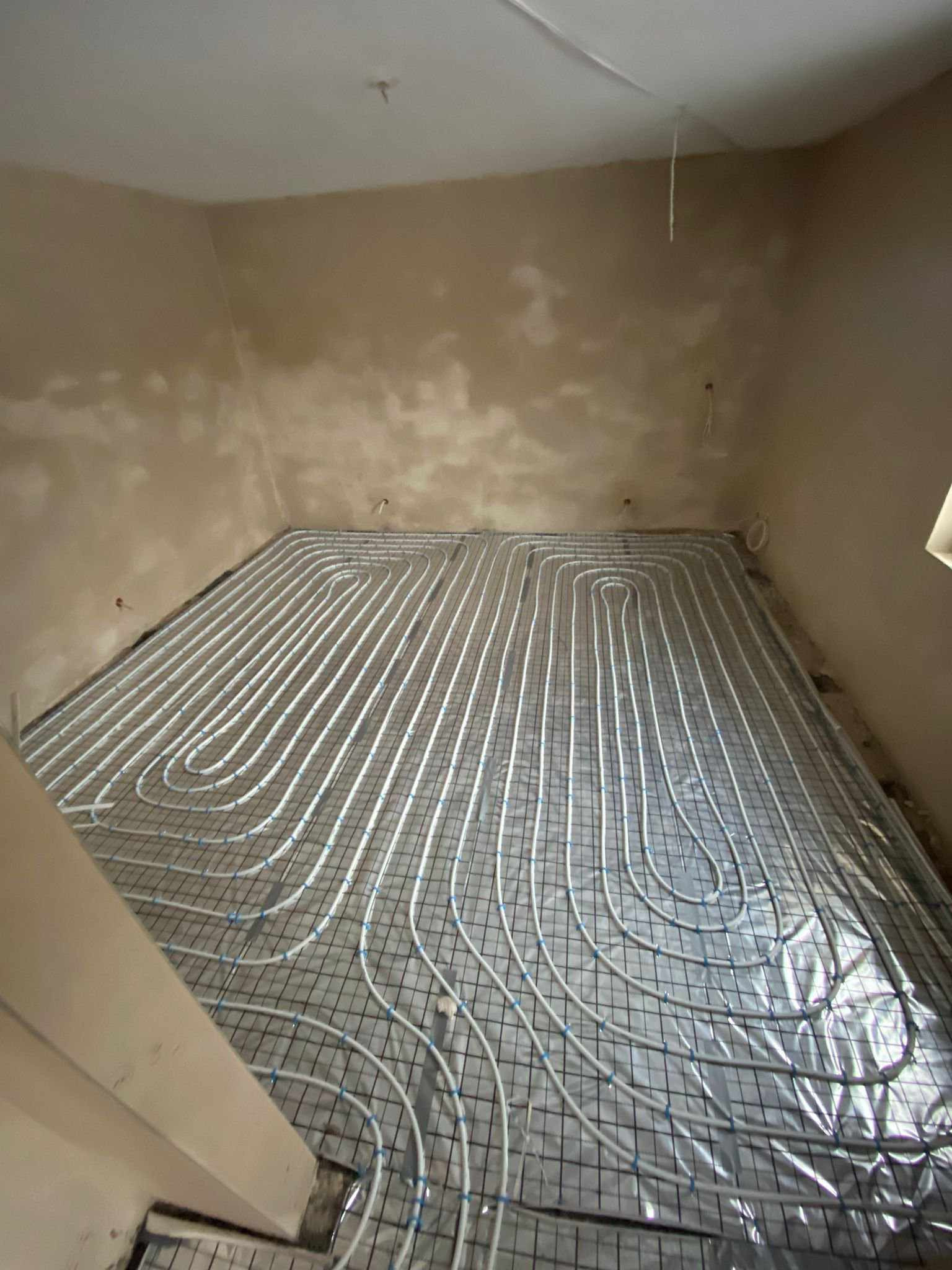 Białe rurki ogrzewania podłogowego ułożone na metalowej siatce na folii izolacyjnej w surowym pomieszczeniu przed wylewką.