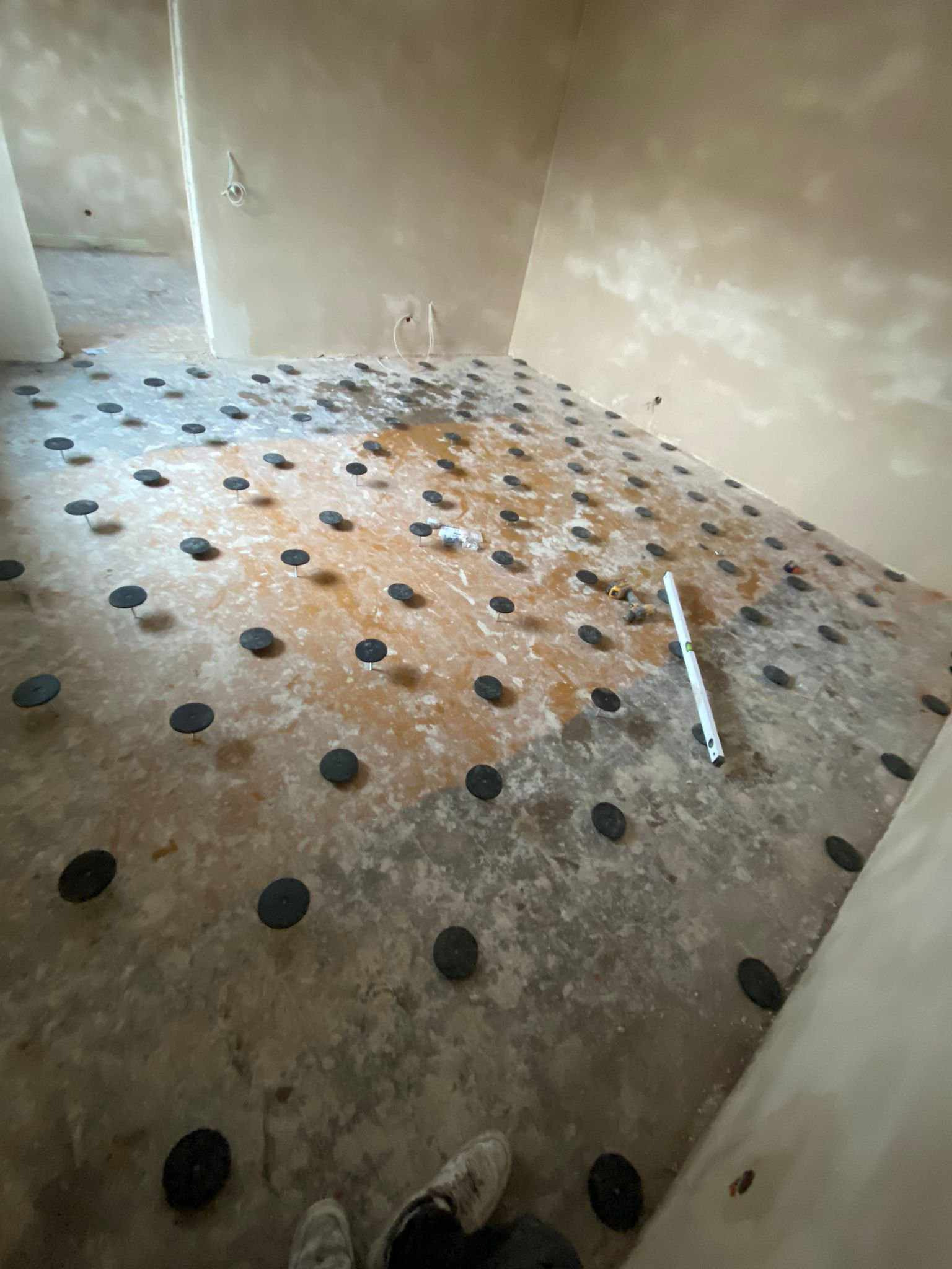 Przygotowanie podłoża pod ogrzewanie podłogowe z widocznymi czarnymi podkładkami poziomującymi rozmieszczonymi na surowej wylewce, poziomica leżąca obok, surowe ściany w tle.