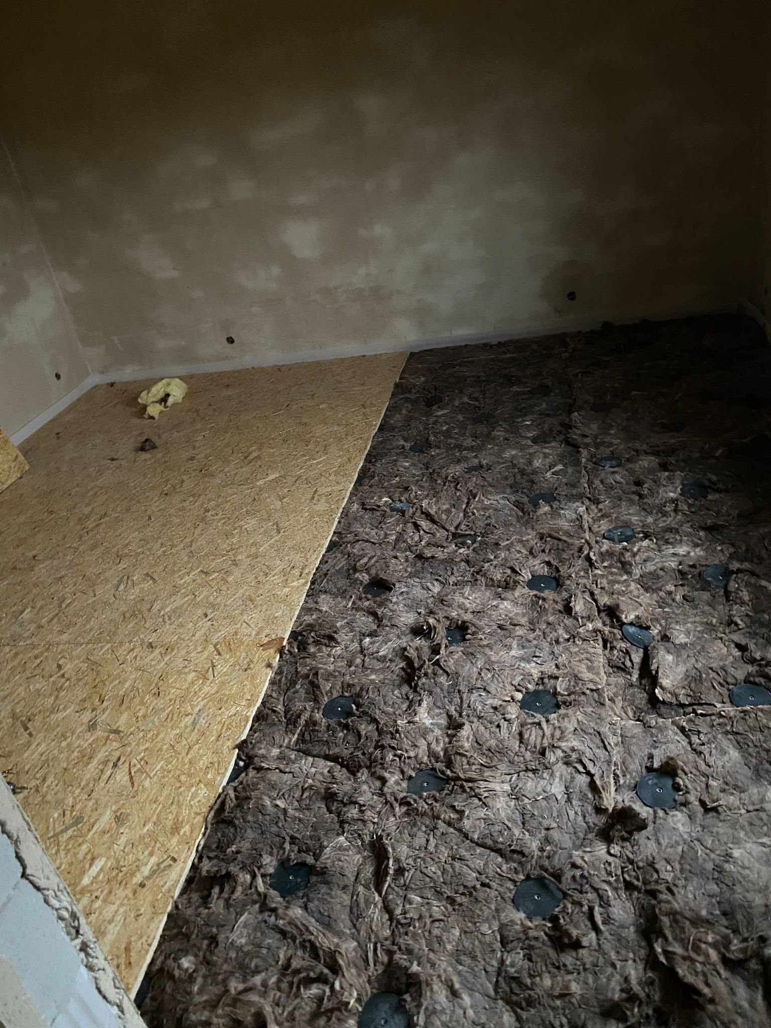 Fragment podłogi w trakcie montażu: część pokryta izolacją z czarnymi elementami mocującymi, a część płytą OSB. Widoczne surowe ściany i narożnik pomieszczenia.