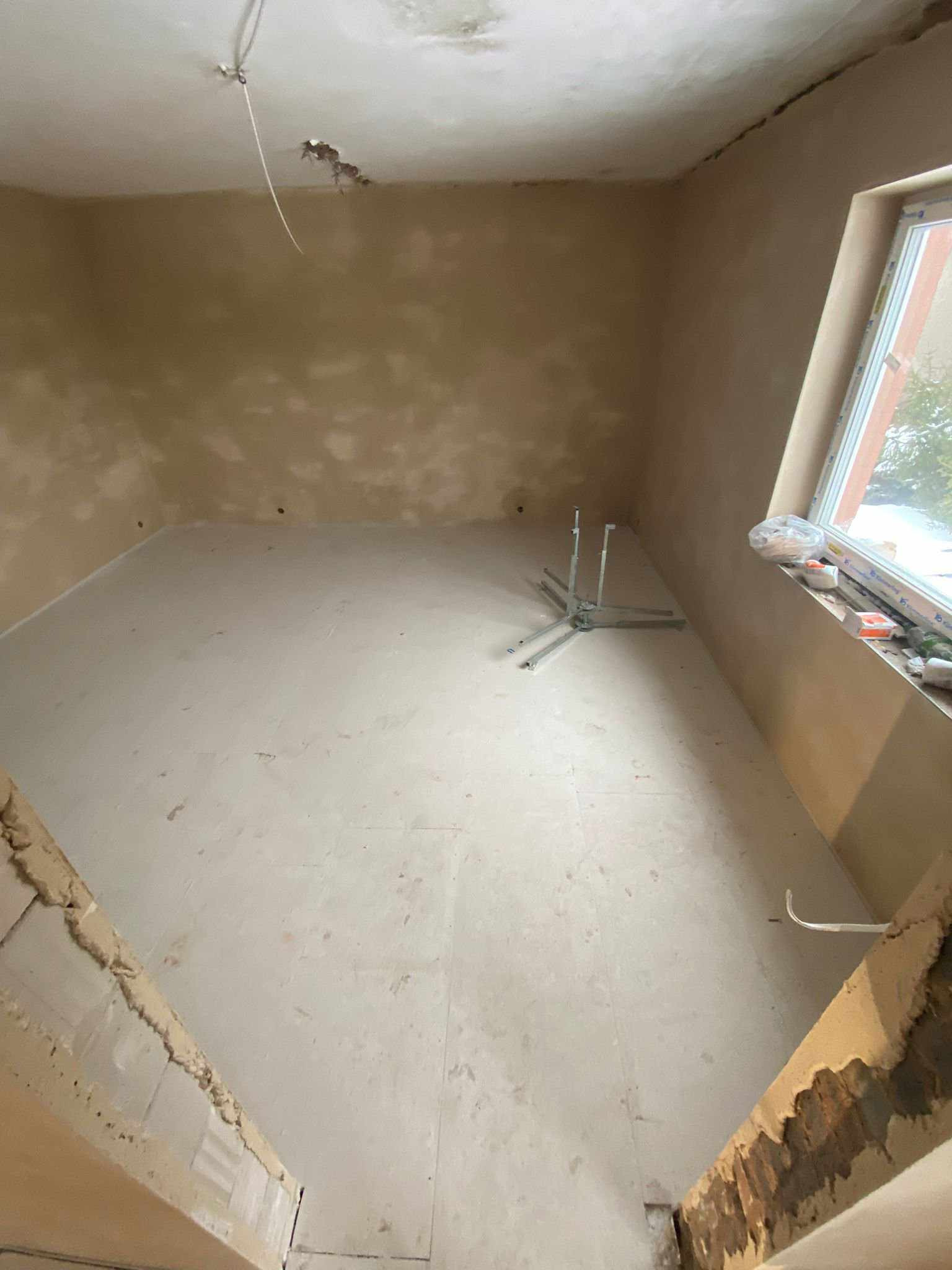 Surowe wnętrze w trakcie remontu: świeżo wylana wylewka podłogowa, tynkowane ściany z widocznymi niedoskonałościami, metalowy stojak na środku pomieszczenia, okno z widocznymi elementami...